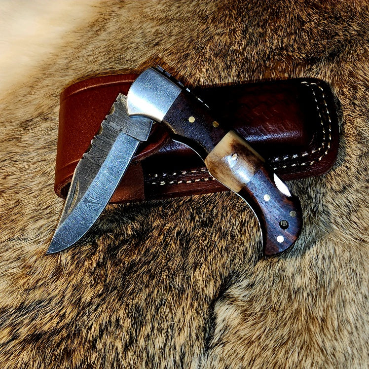 The Walnut Wood & Stag Damascus steel folding knife from Rugged Edge Blades laying on its hand-stitched leather sheath.