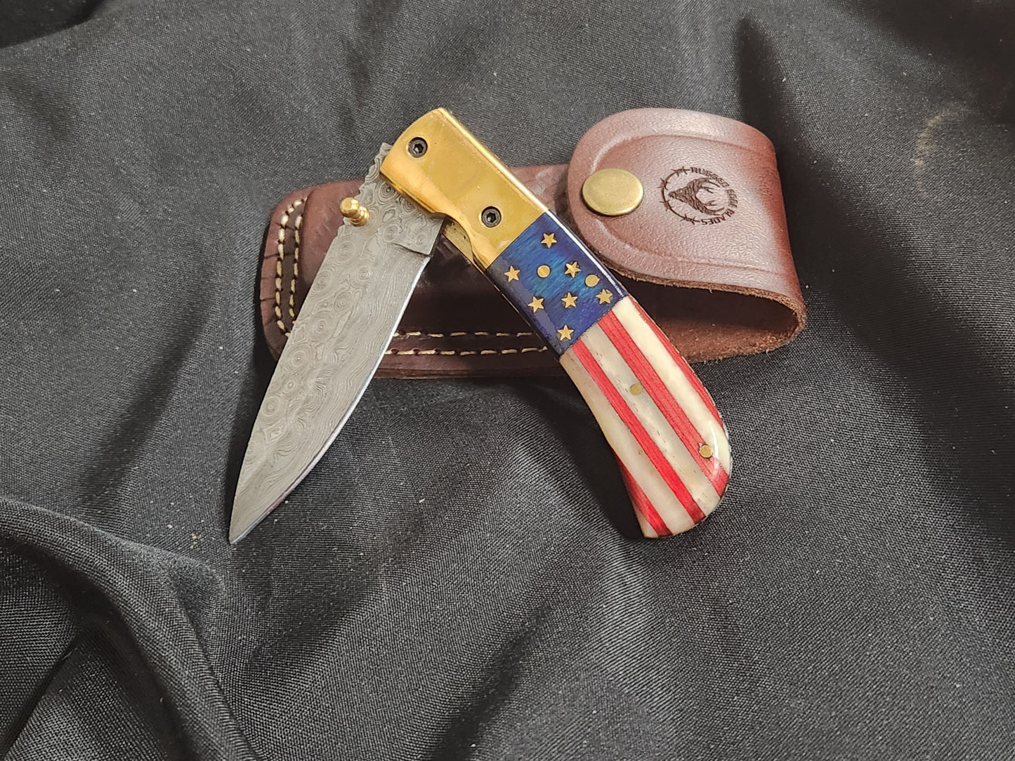 Large American Folder custom Damascus knife with brass bolster and American flag handle, displayed with leather sheath on black fabric.