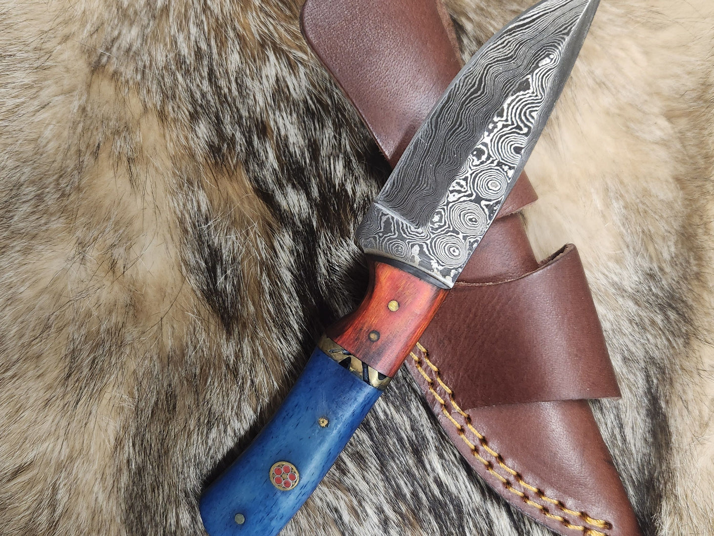Mini Garnet Starlight knife with Damascus steel blade and red-blue resin handle, displayed on fur with brown leather sheath. Ideal for camping and survival use.