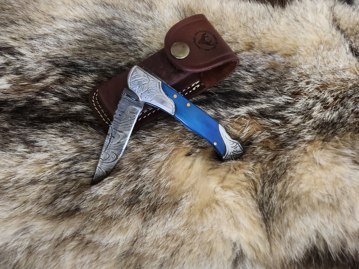 Slim Dyed Bone Blue Folder knife with Damascus steel blade and vibrant blue bone handle, displayed on fur with leather sheath for outdoor use.