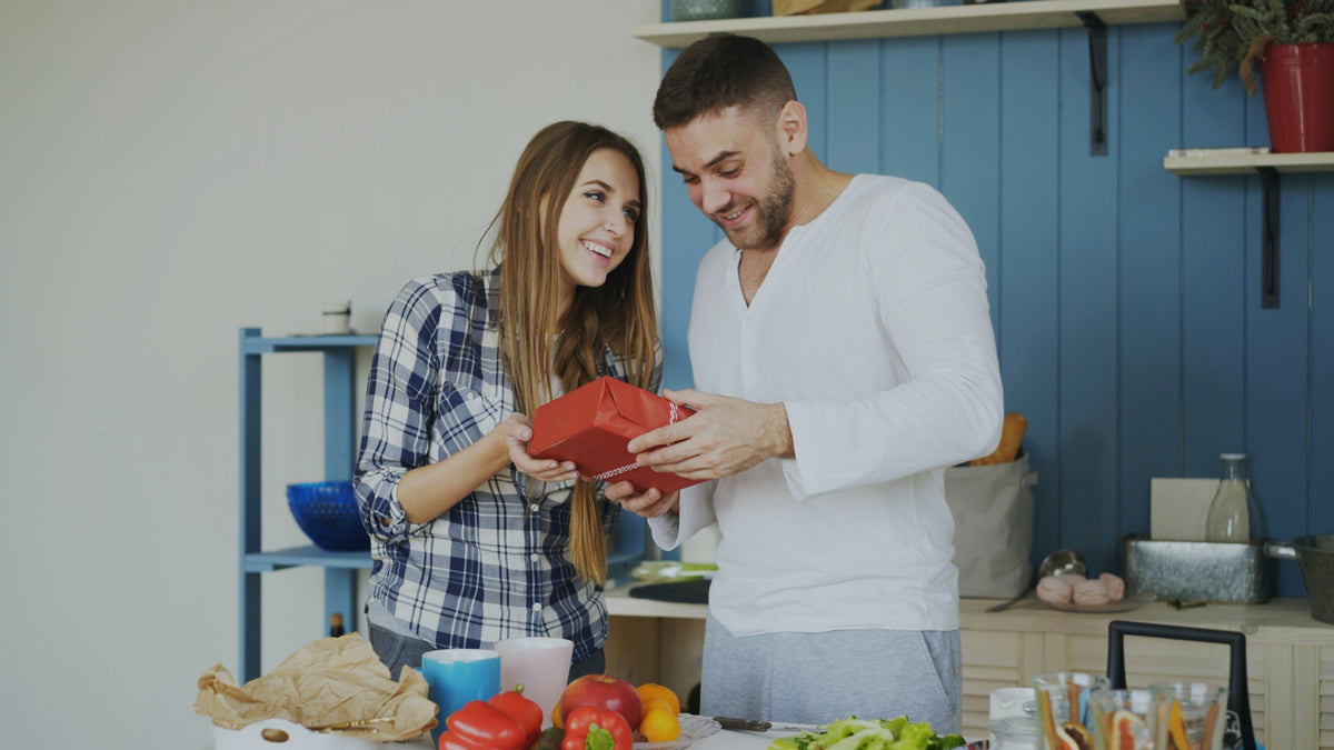 A man receiving a red wrapped gift from a woman while standing in his kitchen.