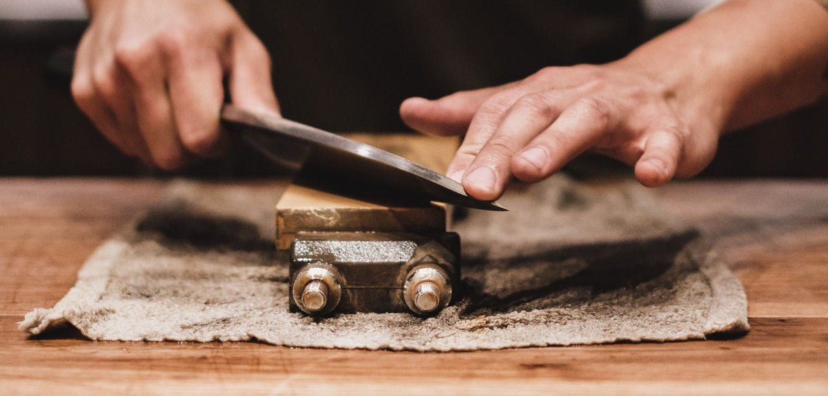 A man in a black t-shirt sharpening his kitchen knife using a whetstone on a kitchen counter.