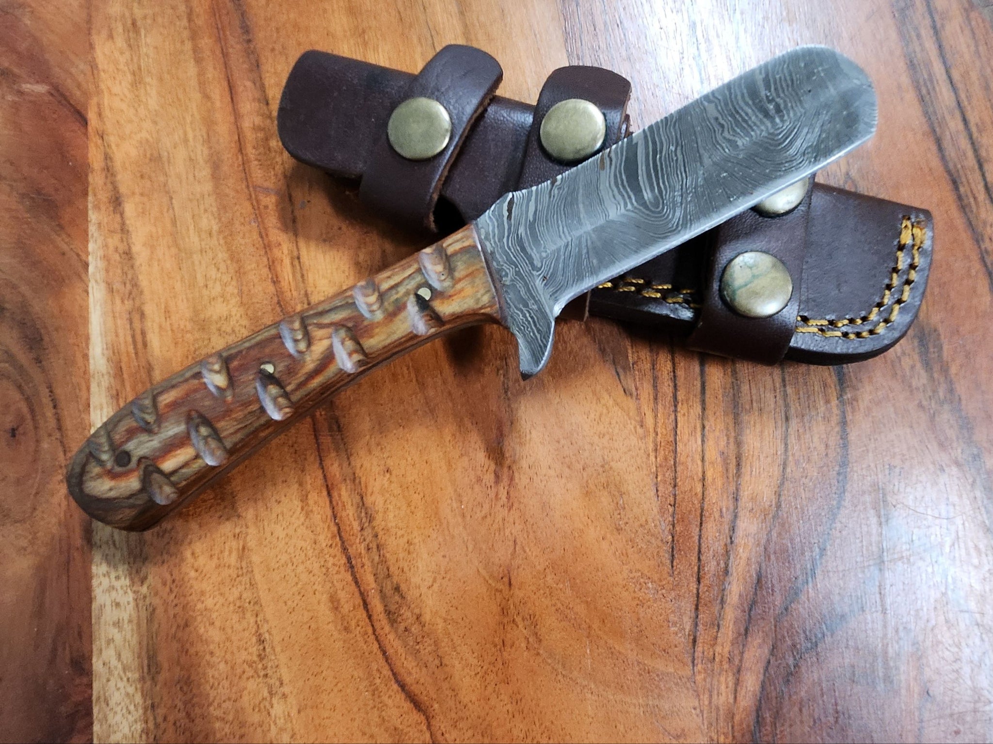 Carved Oak Bull Knife with a Damascus-style blade and intricately carved oak handle, displayed on a leather sheath over a wooden surface.