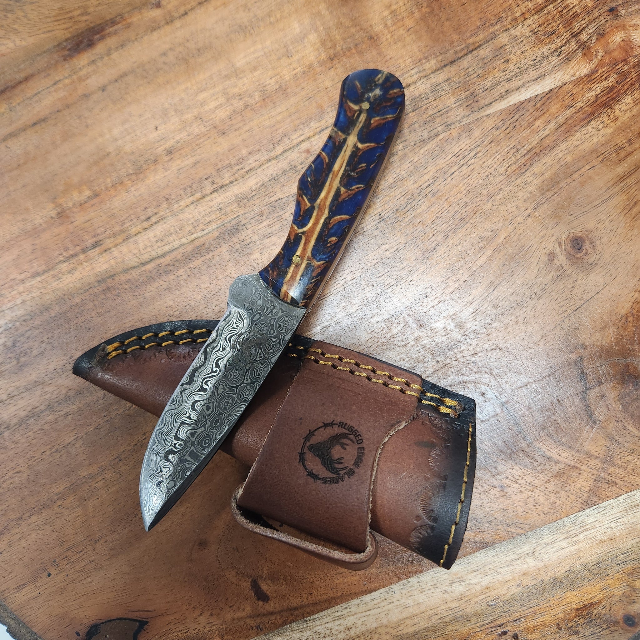 Blue Pinecone EDC fixed blade knife with patterned Damascus steel blade and ergonomic blue resin handle, displayed on a rustic leather sheath.