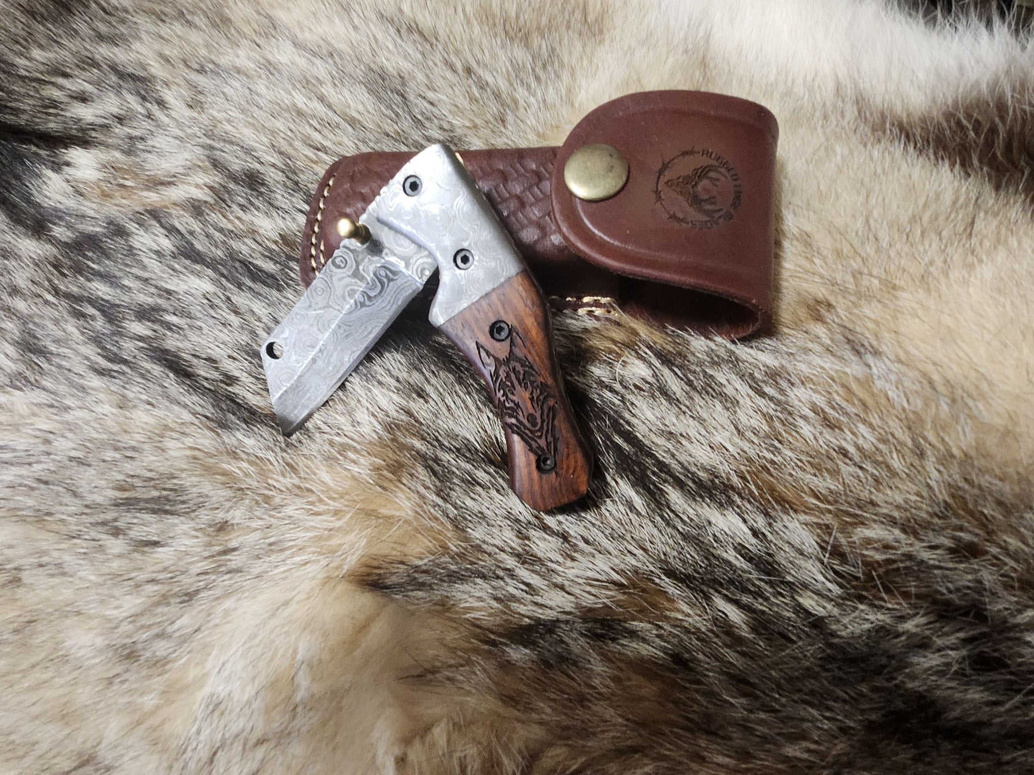 Walnut Wolf Folder knife with Damascus steel blade and engraved wolf design on walnut handle, displayed on fur with leather sheath. Ideal for collectors.