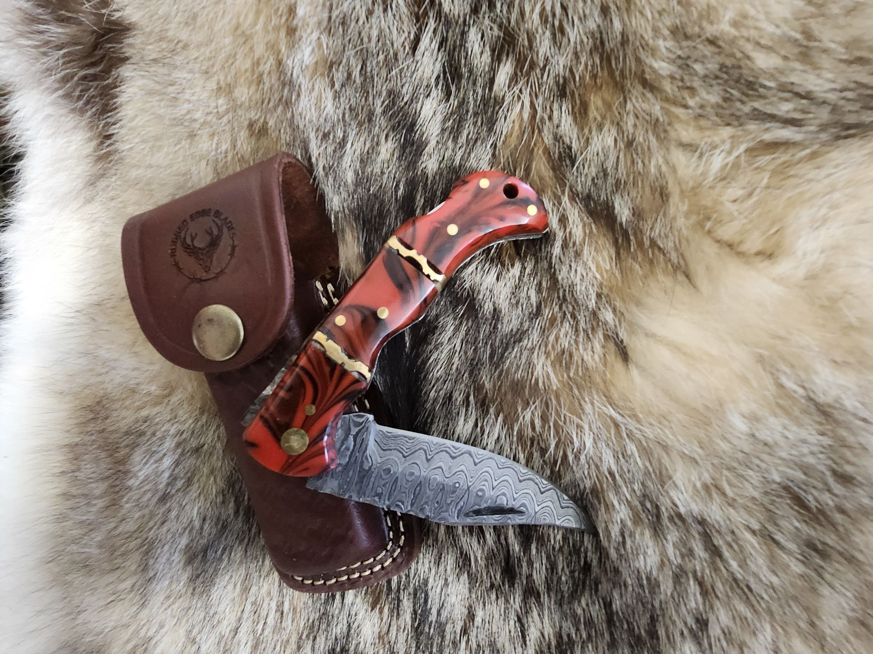 Red Resin Folder knife with Damascus steel blade and brass pins, displayed on fur with a brown leather sheath featuring a stamped logo.