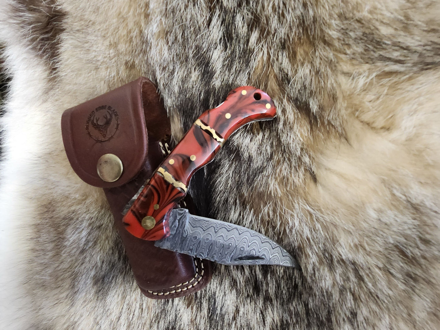 Red Resin Folder knife with Damascus steel blade and brass pins, displayed on fur with a brown leather sheath featuring a stamped logo.