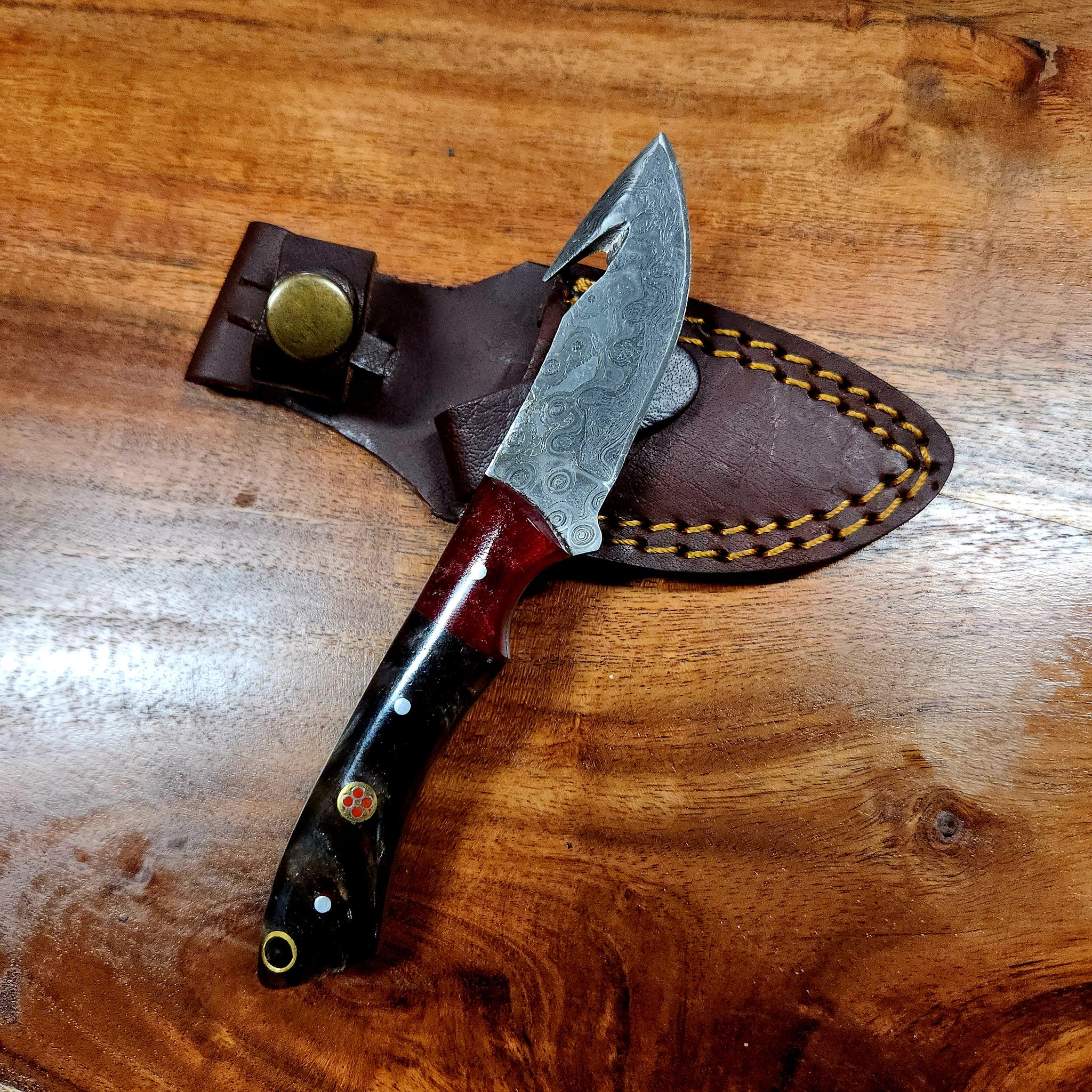 Small Gut-Hook hunting knife with Damascus steel blade and ergonomic handle, displayed with brown leather sheath on wooden surface.