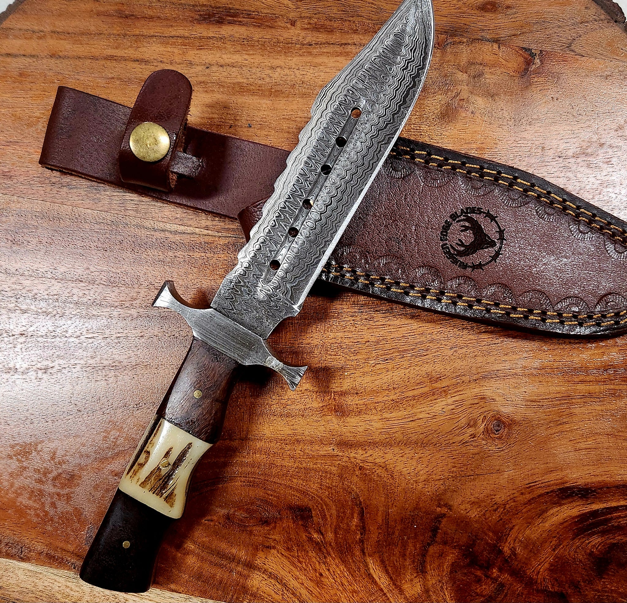 Hunter Bowie knife with a Damascus steel blade and antler handle, displayed with a custom leather sheath on a wooden surface.